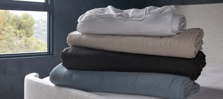A stack of quilts and blankets in white, tan, dark grey, and soft blue on top of a bare Parachute mattress