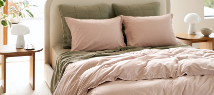 A bed made with moss green and dusty greyish purple cotton percale sheets in a sunny bedroom