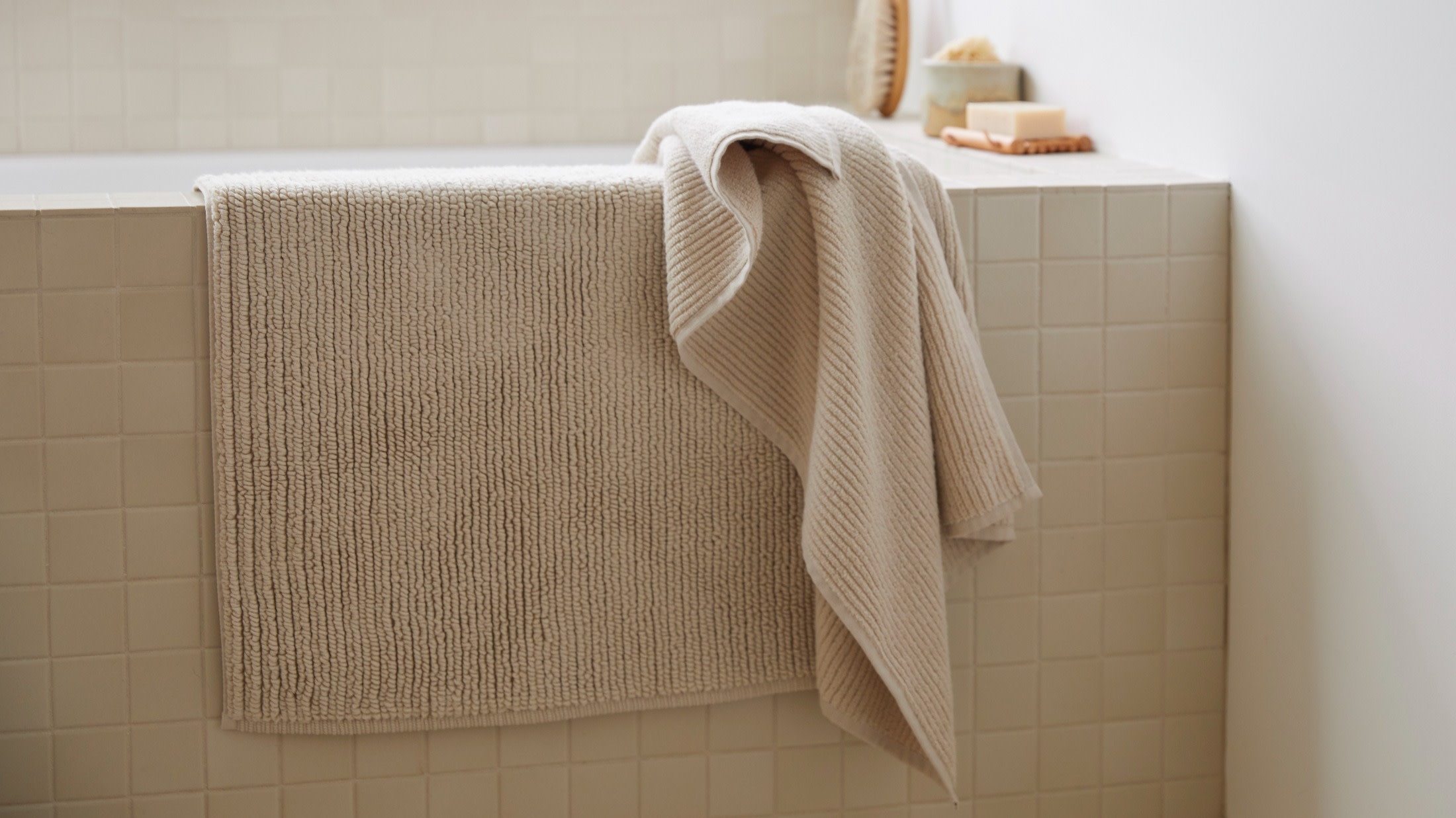 A bone Soft Rib Tub Mat and Bath Towel hanging over the side of a tiled bath tub