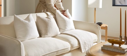 A person fluffing a pillow on a plush, ivory sofa