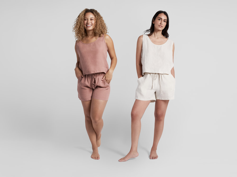 Two women standing on a blank white set wearing linen tanks and shorts.