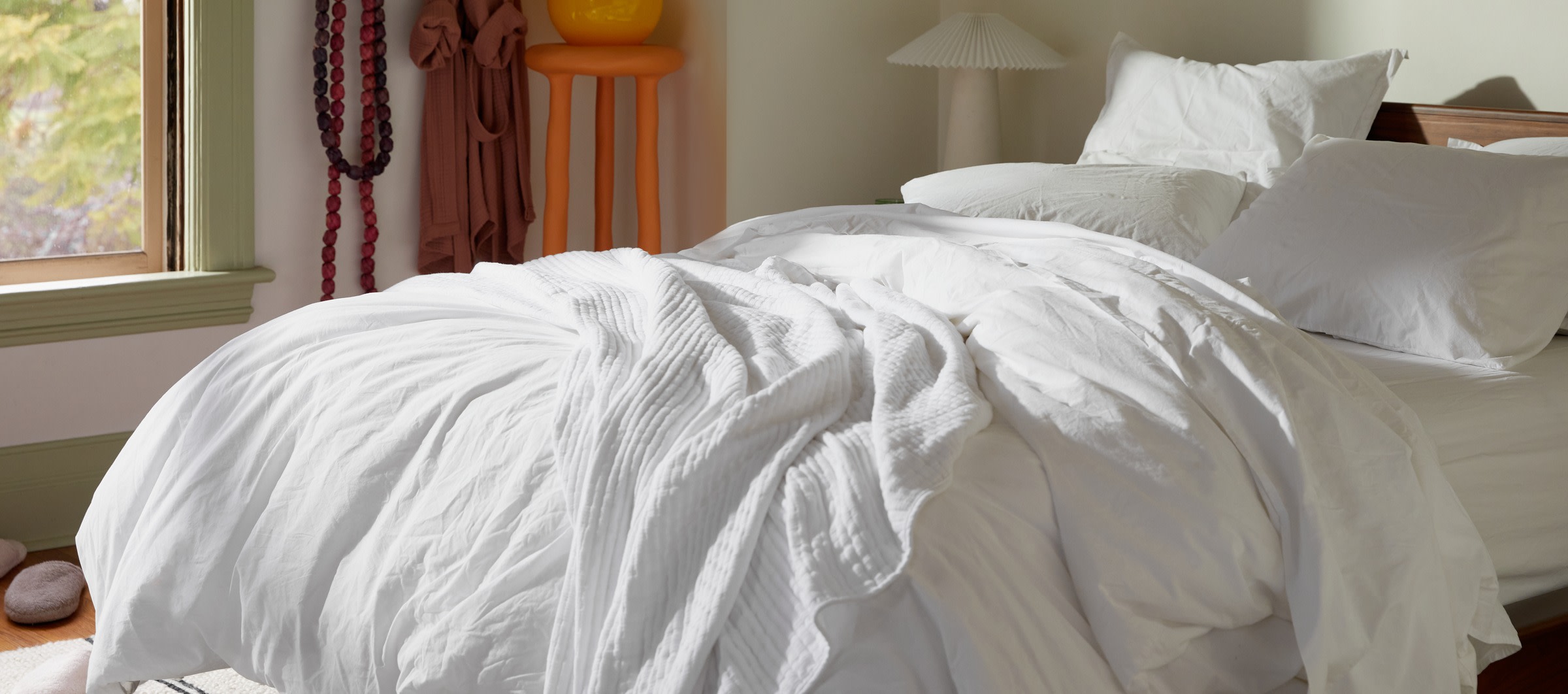 A messy bed with white cotton percale sheets in a room filled with bright, eclectic decor