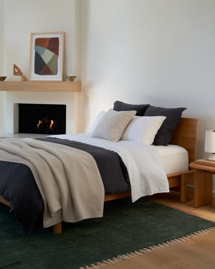 A neatly made bed with dark grey bedding in a bright bedroom with a dark evergreen wool rug