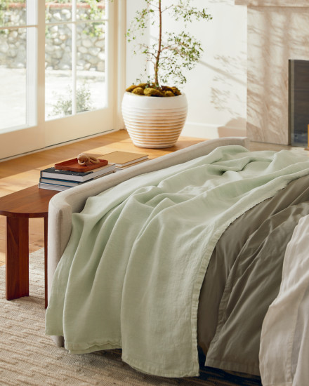 A naturally lit room showcasing a bed frame with a mixture of bedding with a wooden table at the foot of the bed.