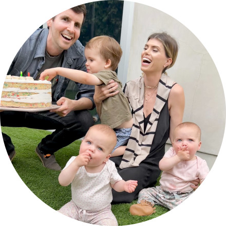 A photo of a smiling woman holding three babies and her partner smiling and holding a birthday cake