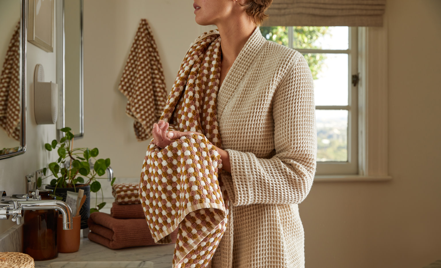 Woman standing in a bathroom wearing a tan waffle robe wiping her hands with a mosaic hand towel