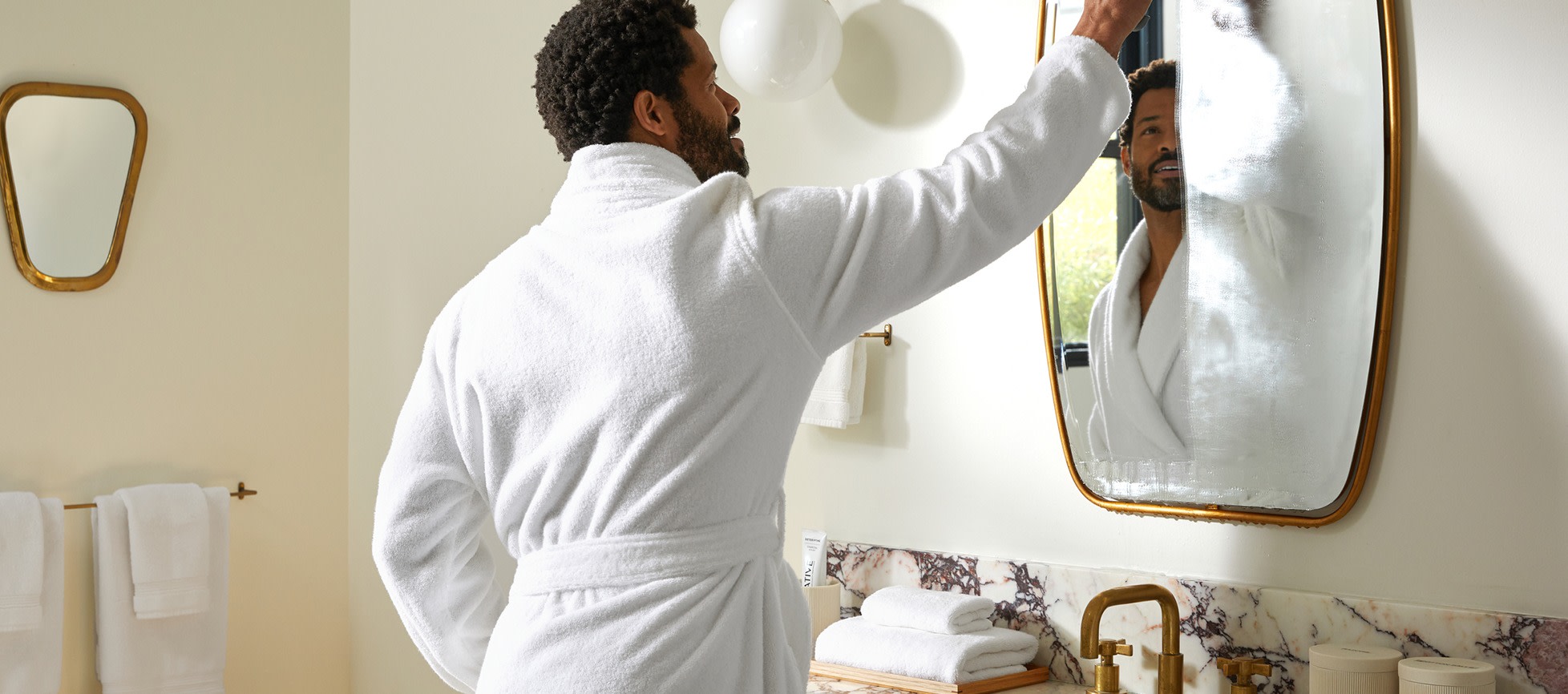A man wearing a plush white bathrobe and wiping a foggy mirror