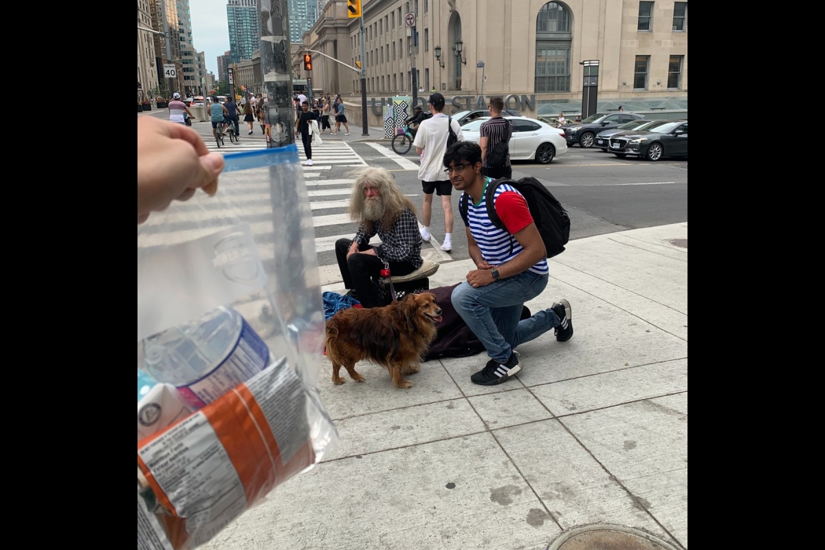 A homeless individual and their dog are seen at a street corner in front of Toronto Union Station. A member of Blankets for T.O. is seen kneeling next to him.