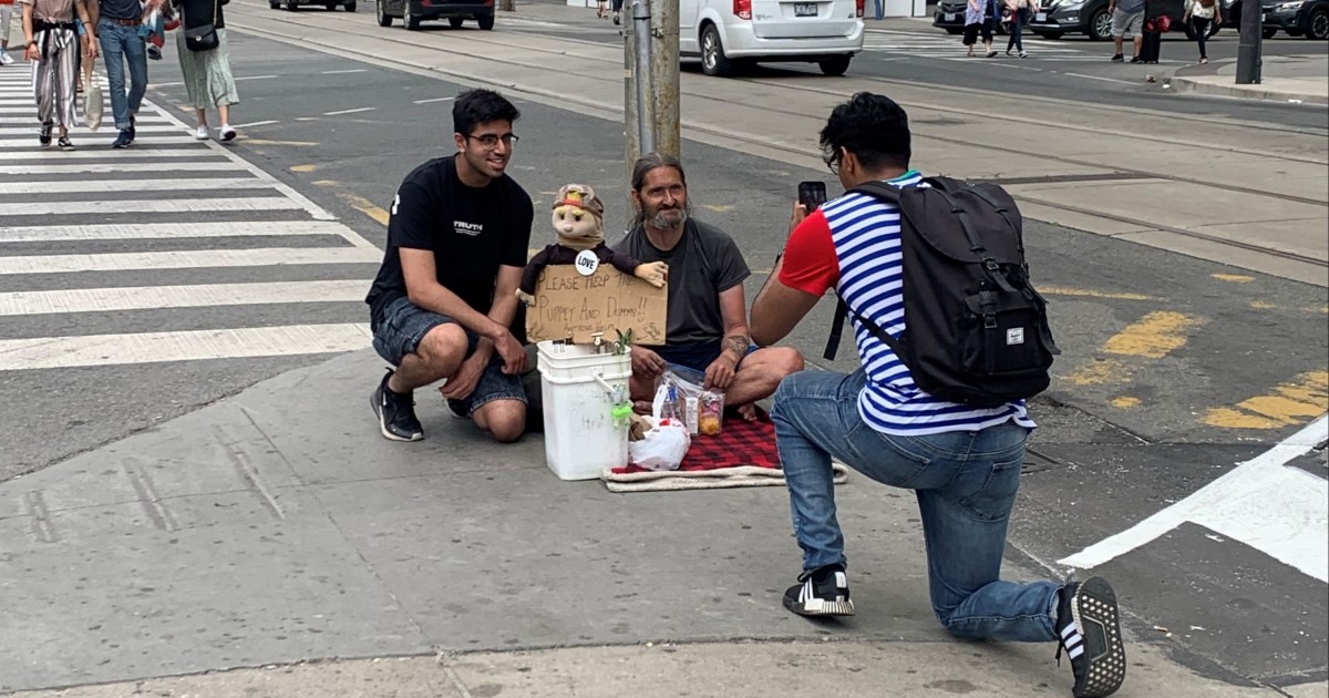A Blankets for T.O. executive team member takes a picture of another donation run participant posing with a homeless person sitting on the sidewalk. The homeless person has a puppet and sign.