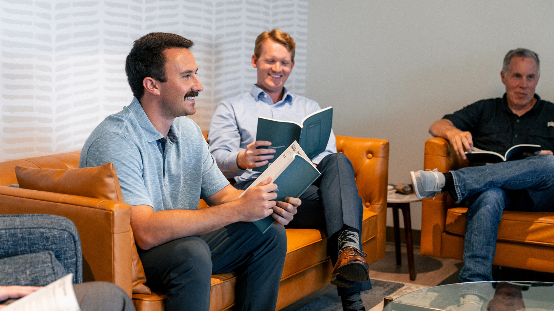 Men in a small group participating in the Christian discipleship care & recovery ministry of Re:generation, sitting on leather couches, reading and discussing the ministry curriculum in a modern, comfortable setting.