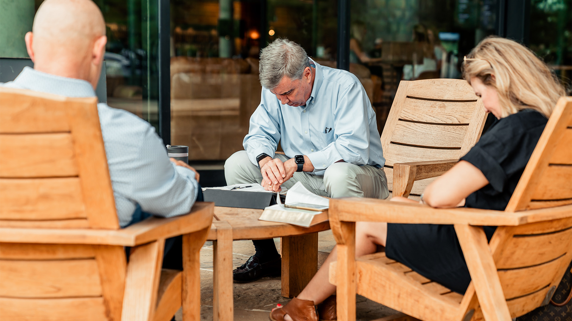 A group of three people praying outdoors on wooden chairs at Watermark Community Church in Dallas, Texas, with Bibles and notebooks on a table, engaged in spiritual reflection.
