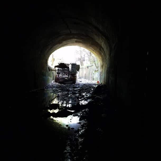 Inside a dark train tunnel, with puddles reflecting light. A rusty crane is partially illuminated at the entrance, framed by sunlight.