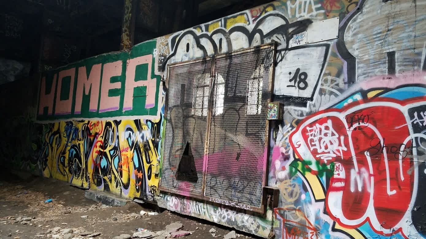 Tunnel with extensive, vibrant graffiti. Chain-link metal gate in centre, dark interior behind. Debris on the ground.