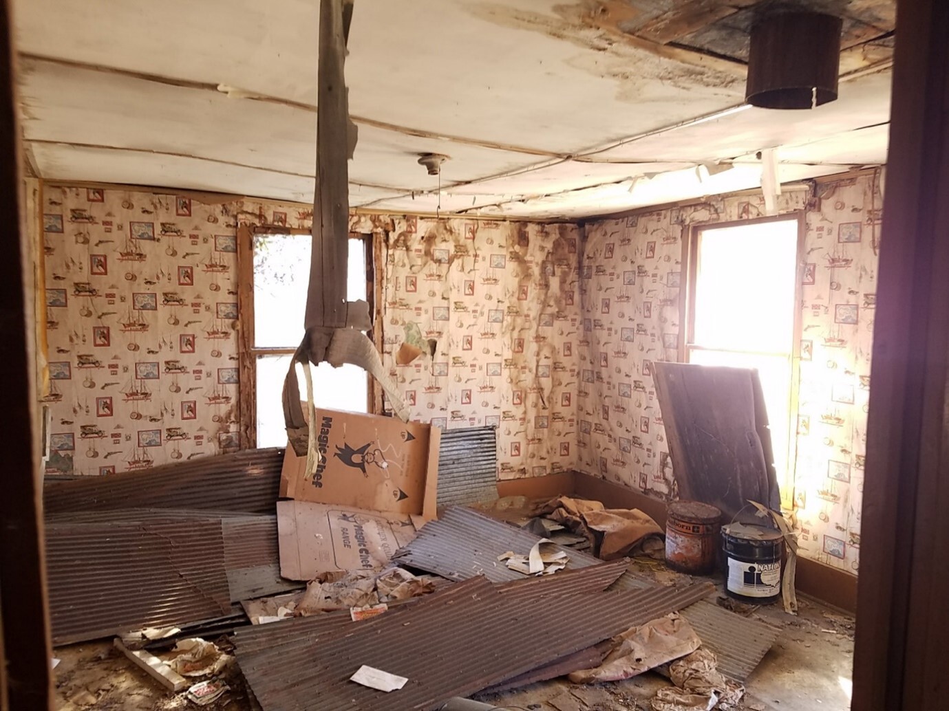 An abandoned room in a house with peeling wallpaper, debris, and metal sheets on floor. Sunlight filters through windows.