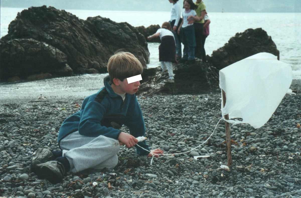 Pictured: “Potential Juvenile Incarnation of the Cloud MillionAIRe: A Ten-Year-Old with an Improvized Kite Utilizing a White Plastic Bag on a Coastal Landscape.” A young boy, believed to be the nascent figure of the Cloud MillionAIRe