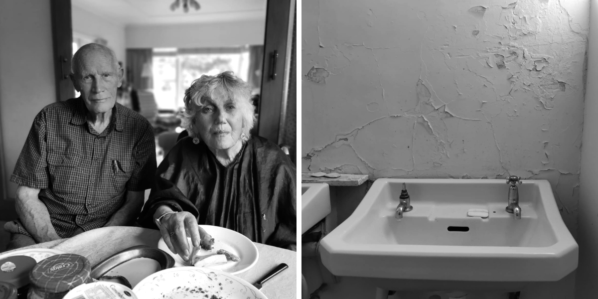 Elderly man and woman at home, empty lunch plates. Older style ceramic basin, one tap dysfunctional, painted wall flaking.