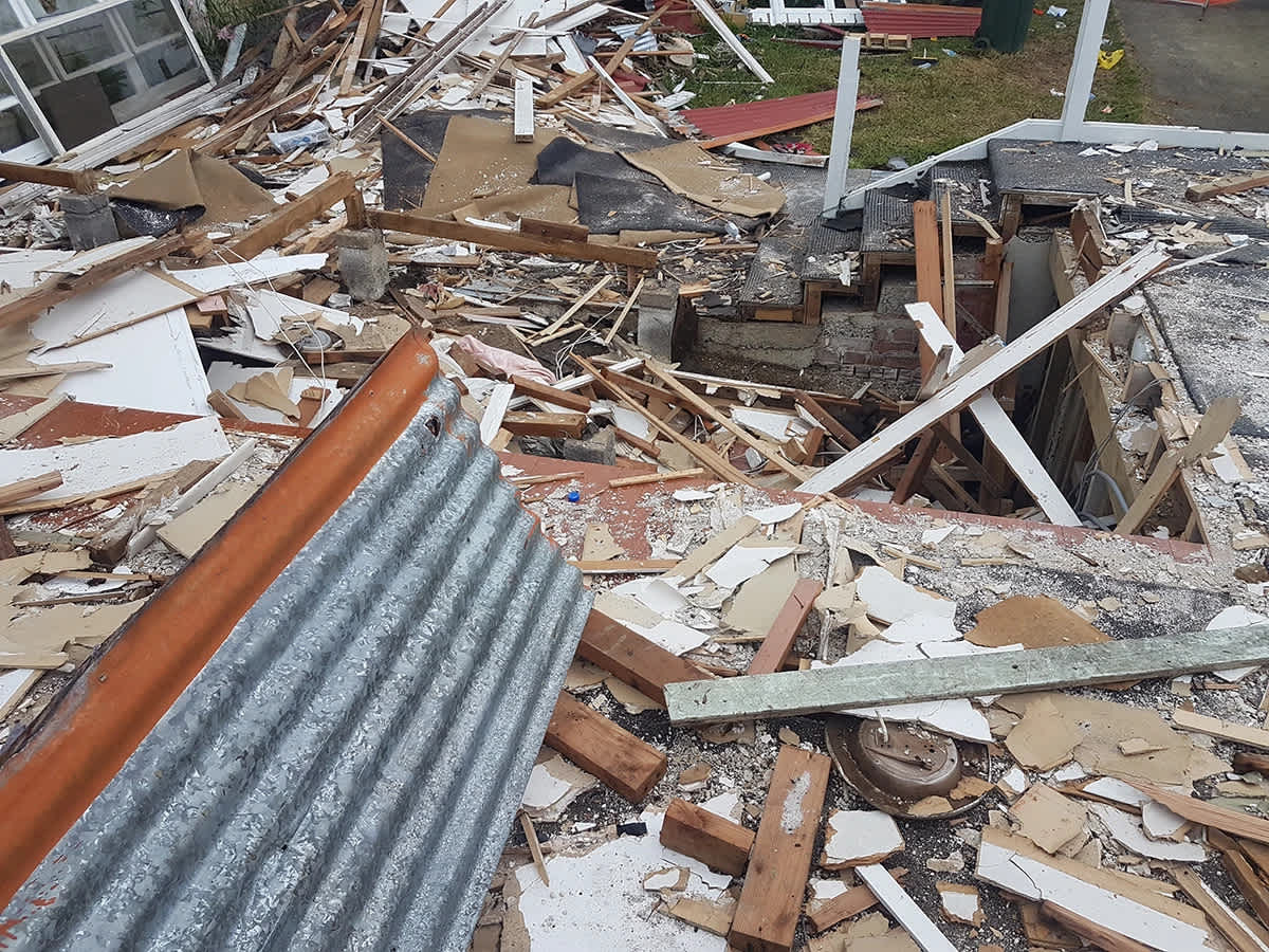 Pictured: “Personal Belongings Amidst Rubble.” A missing window, holes in the ceiling, and ripped-off gib board exposing Pink® Batts® insulation. The bush in the background remains undisturbed.