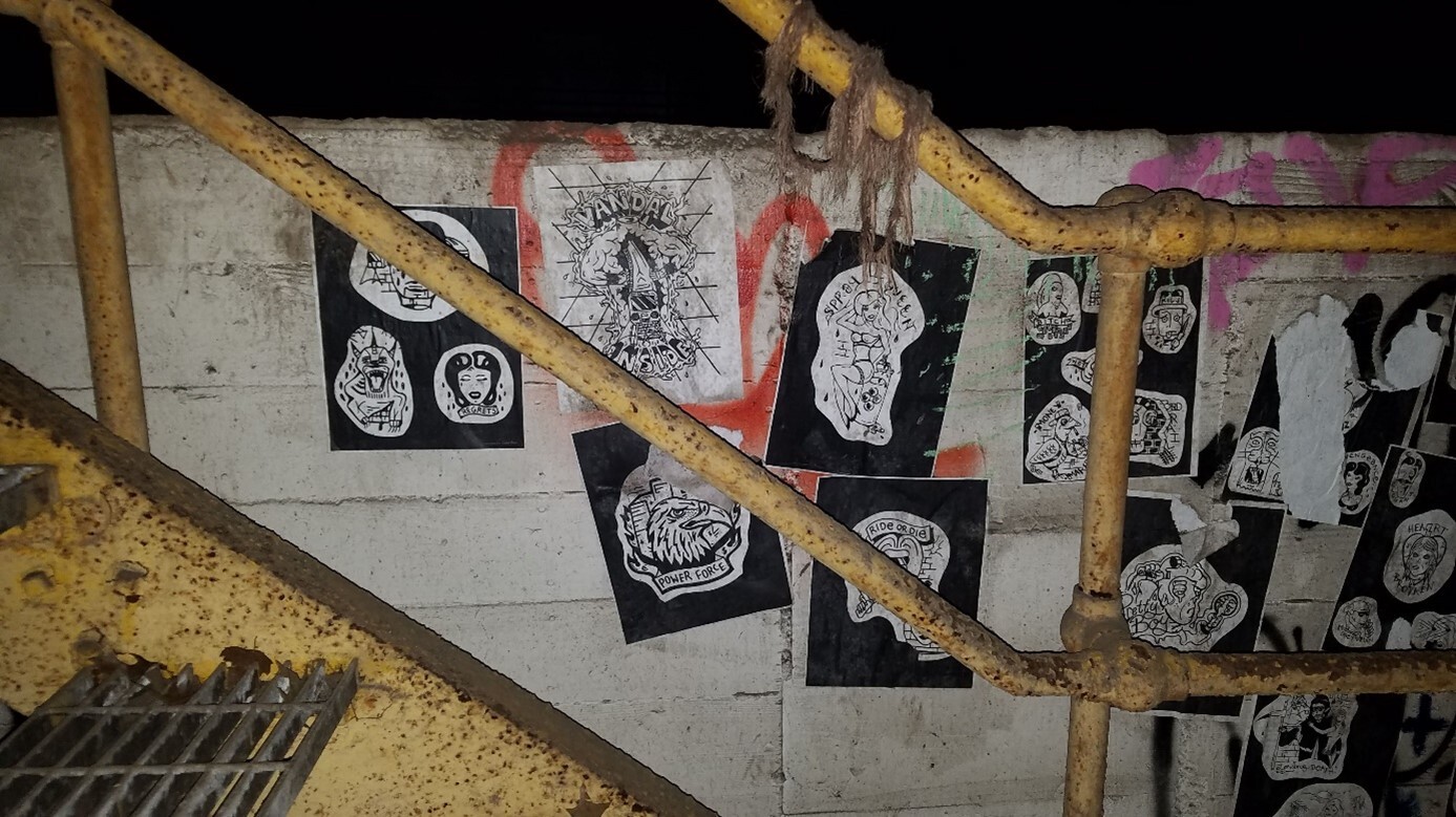 Weathered concrete wall in a train tunnel, covered with black and white posters, graffiti, and a rusty yellow metal staircase railing.