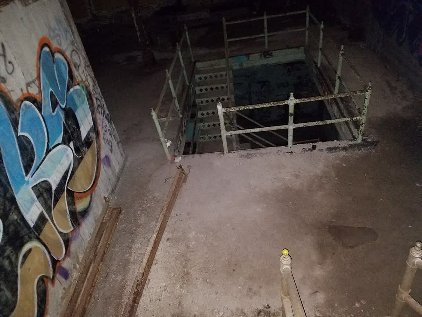 View from above of an abandoned, dirty, cold-storage facility. Metal staircase descends through floor. Graffitied walls.