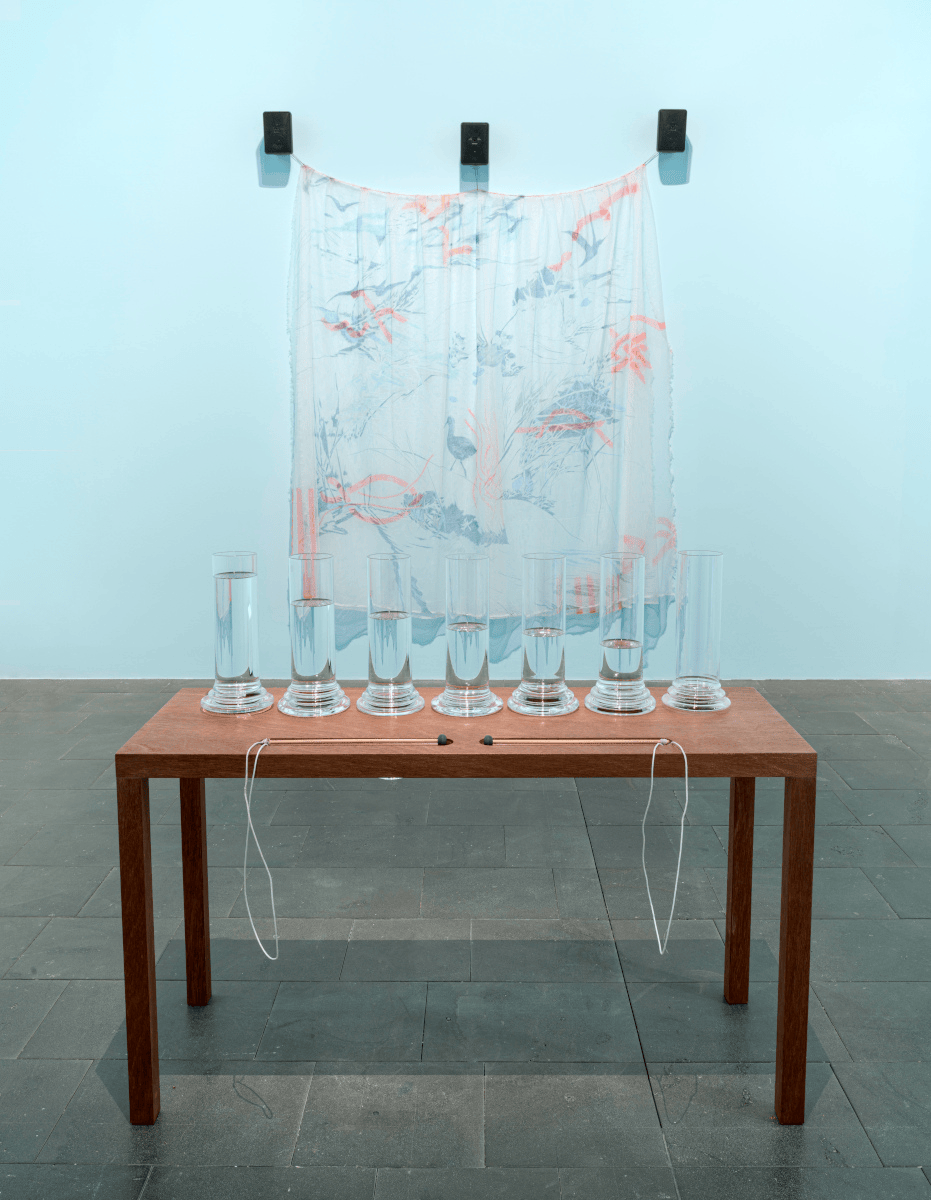 A photo of an instrument made out of glass and water, in front of a drawing suspended from three speakers on a wall.