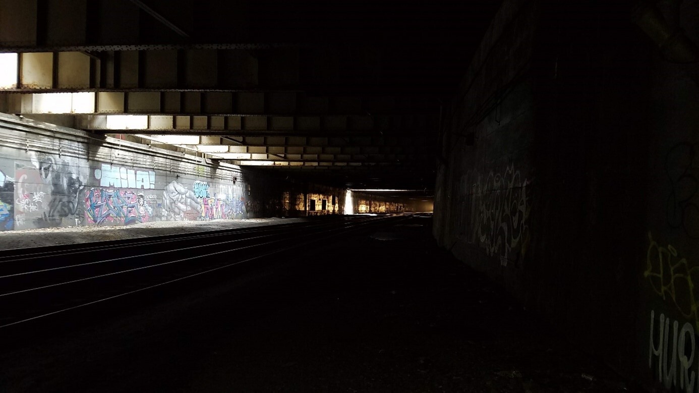 Dimly lit train tunnel with tracks stretching into the distance. Colourful graffiti on the left, distant light illuminating the tracks.