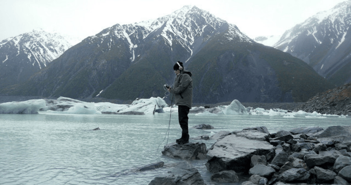 A photo of a person with headphones on and cables in the water stands on a rock in a lake with mountains behind.