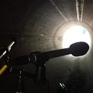 Interior of a metal pipe with two microphones in the foreground. Bright light at the far end contrasts with the dark, textured walls.