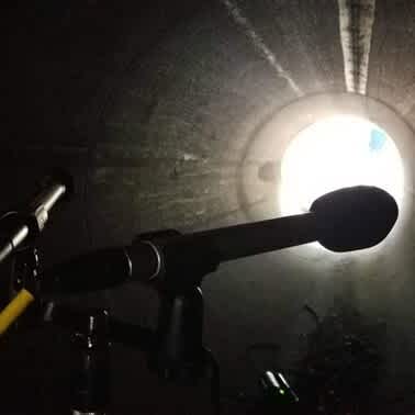 Interior of a metal pipe with two microphones in the foreground. Bright light at the far end contrasts with the dark, textured walls.