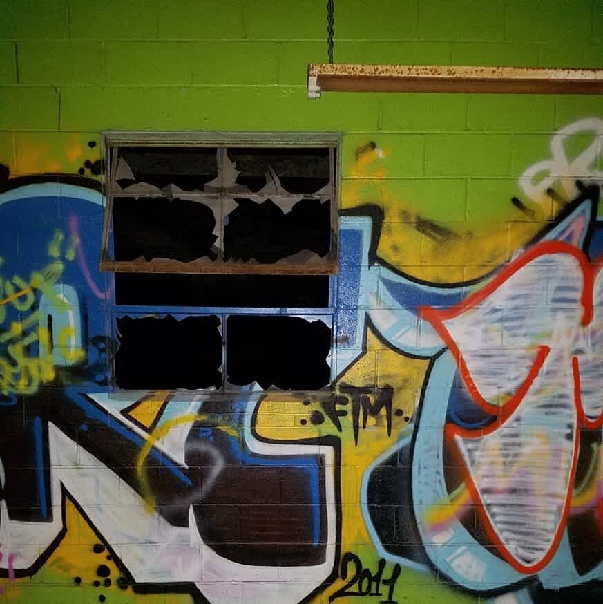 Concrete factory wall painted lime green with graffiti. Open broken window, rusted light fixture hangs by chain.