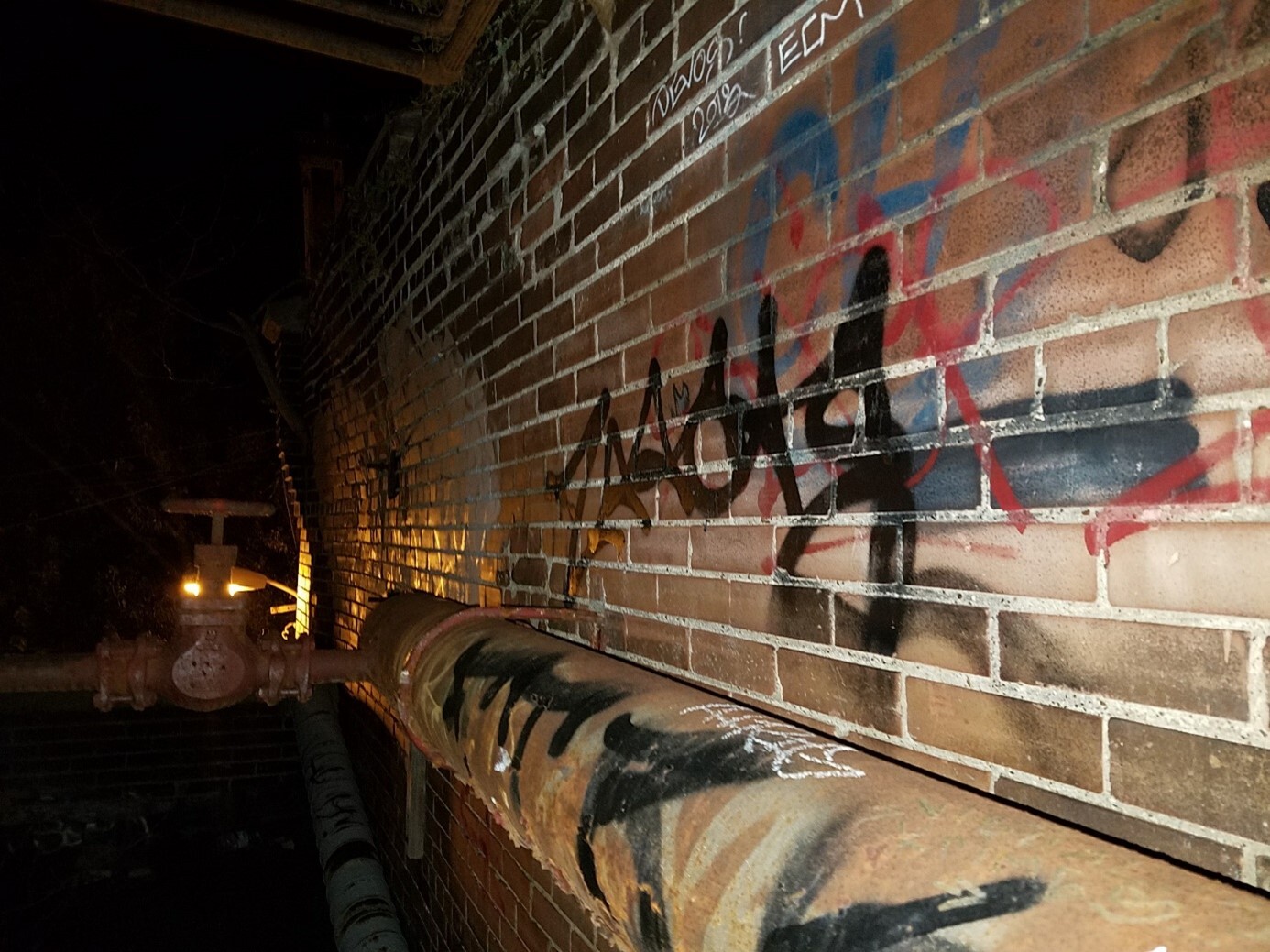 Train tunnel hallway with a brick wall covered in colourful graffiti. A rusty pipe runs parallel to the wall.
