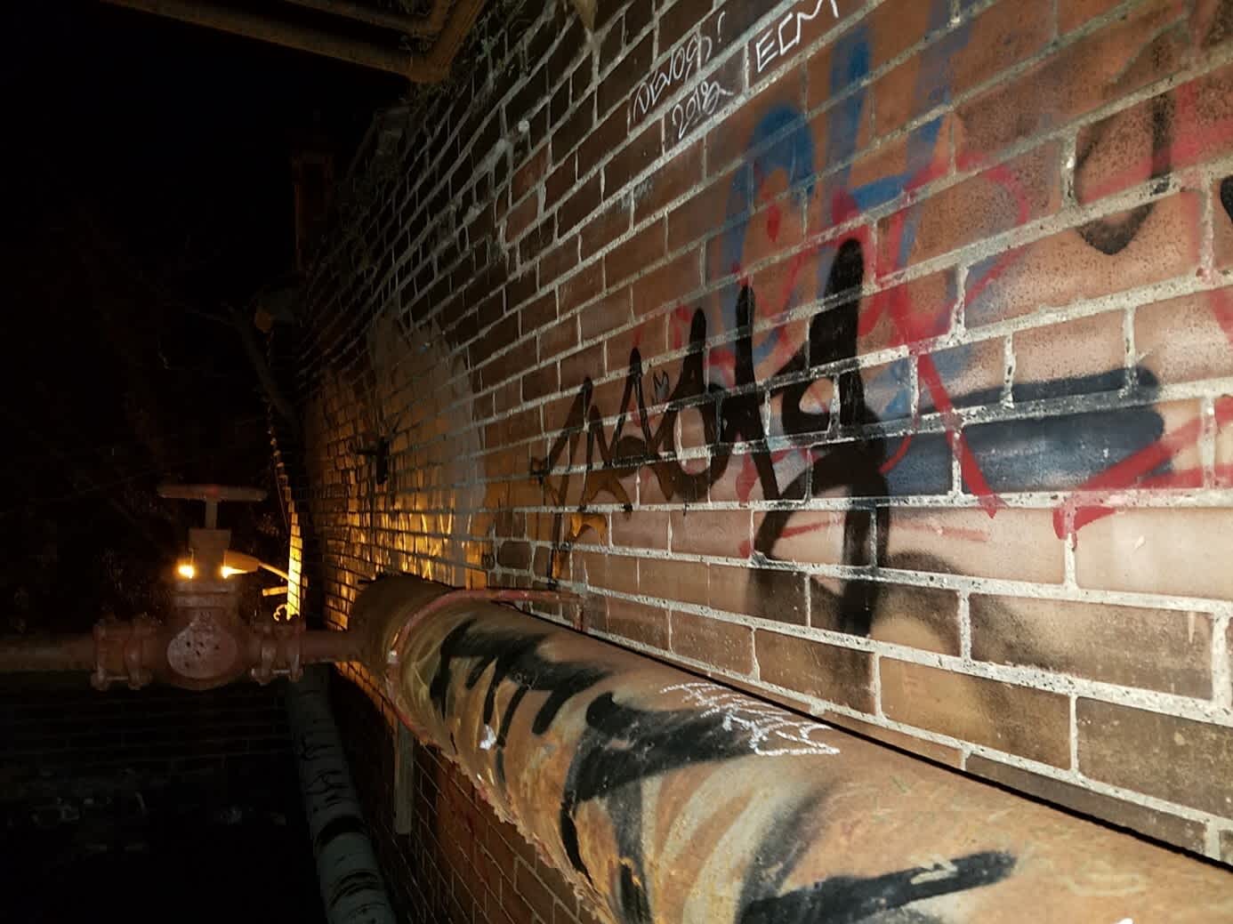 Train tunnel hallway with a brick wall covered in colourful graffiti. A rusty pipe runs parallel to the wall.