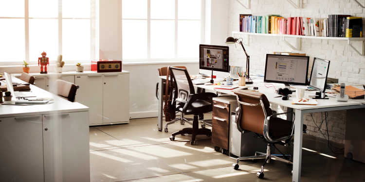 An office with sun coming through larger windows shining on two desks and design supplies.