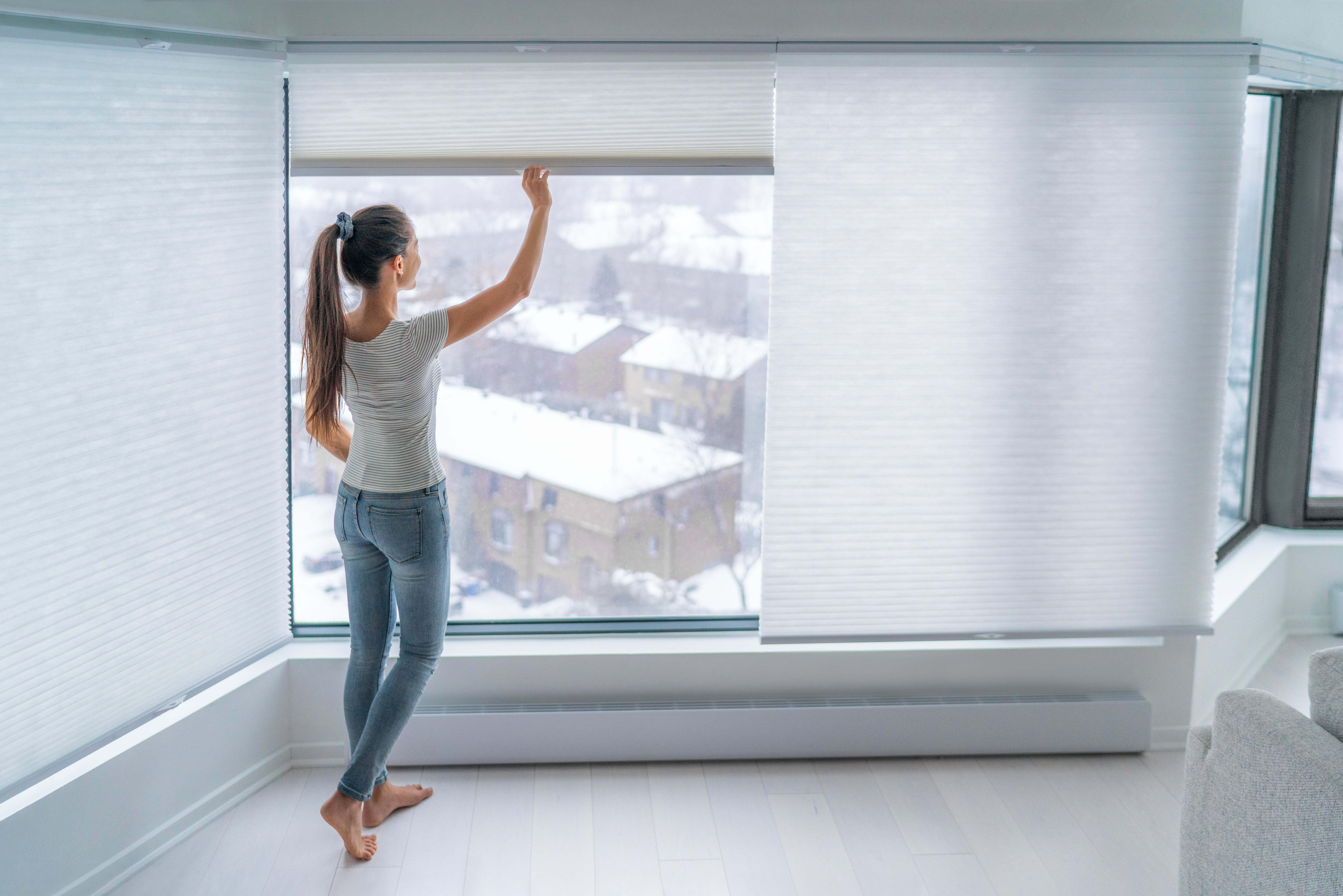 A woman pulling up the blinds to look out a window in her apartment