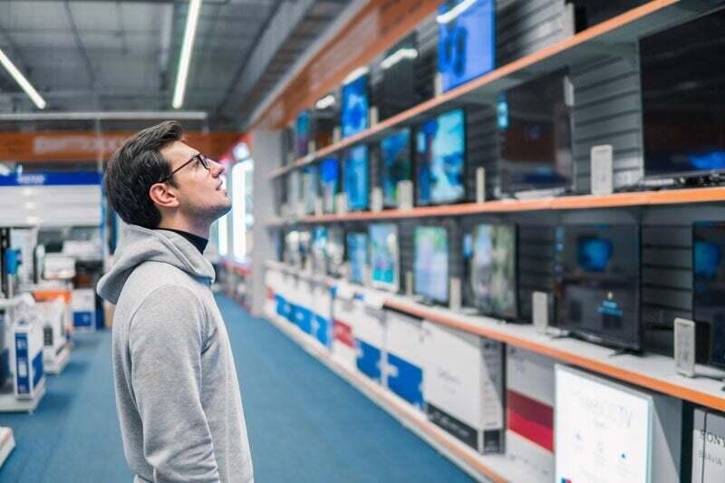 In a large store, a man looks interestedly at shelves with various sizes of televisions.