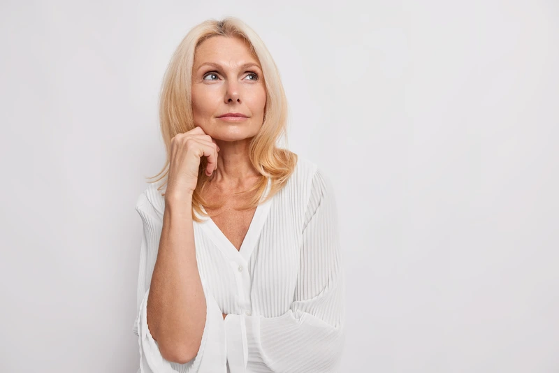 thoughtful-blonde-middle-aged-woman-ponders-something-keeps-hand-near-face-has-healthy-skin-minimal-makeup-makes-choice-wears-white-blouse-poses-indoor-blank-copy-space-your-promotion
