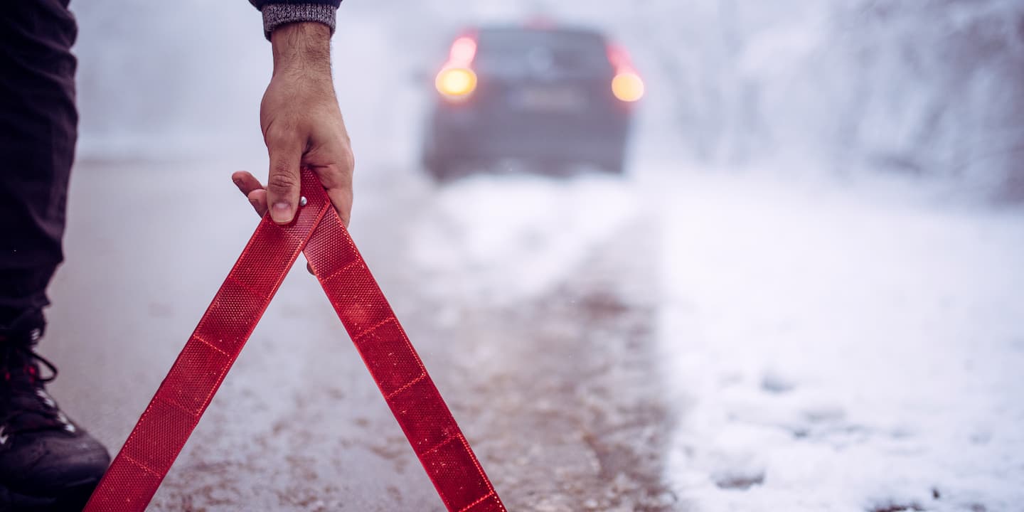 Man placing warning triangle on the road to alert other drivers of his broken down car on a snowy day. 