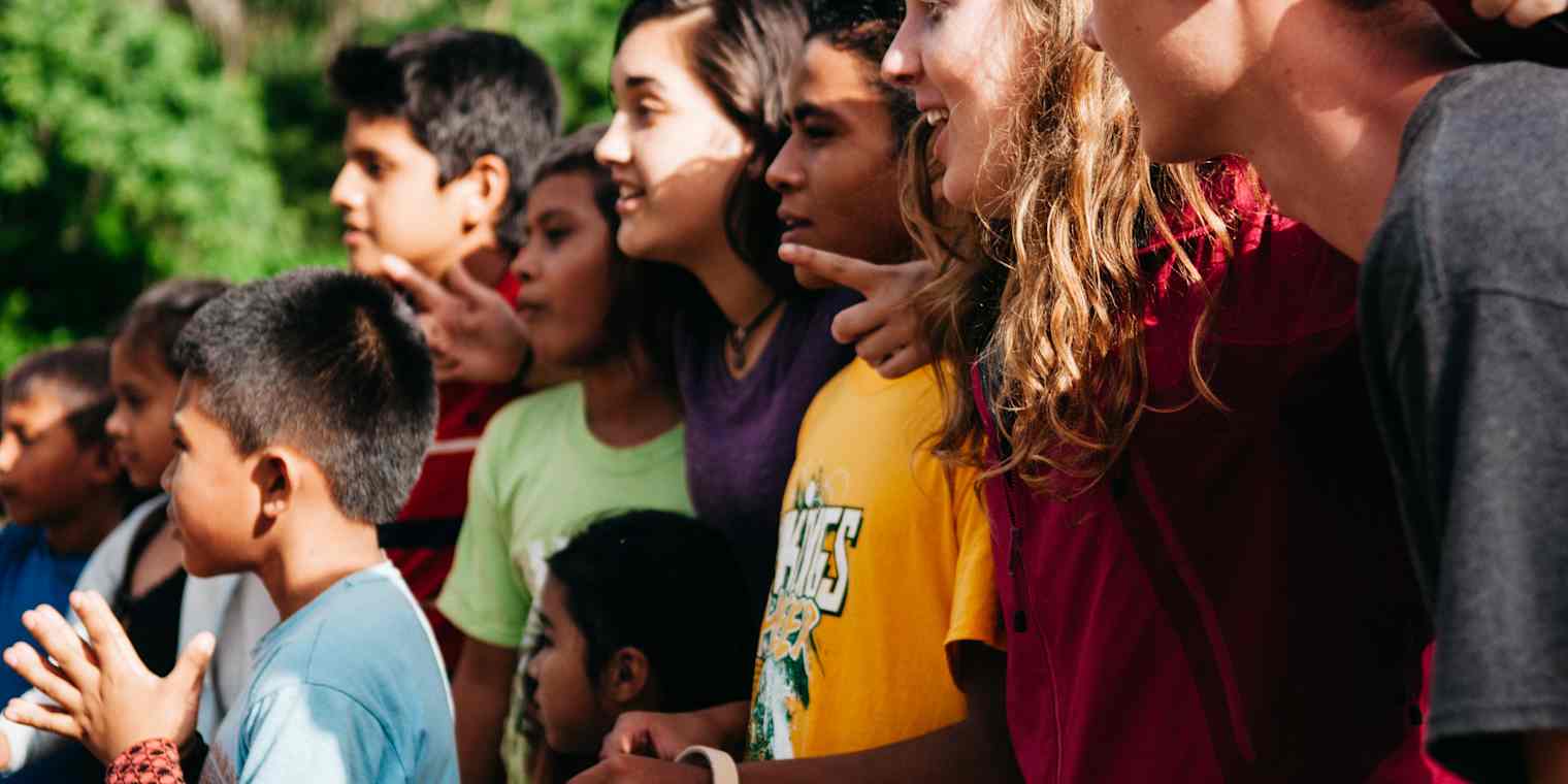 A group of children and young adults stand together, all facing the left. They are smiling and have their arms around each other.