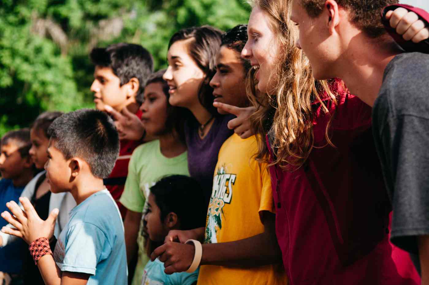 A group of children and young adults stand together, all facing the left. They are smiling and have their arms around each other.