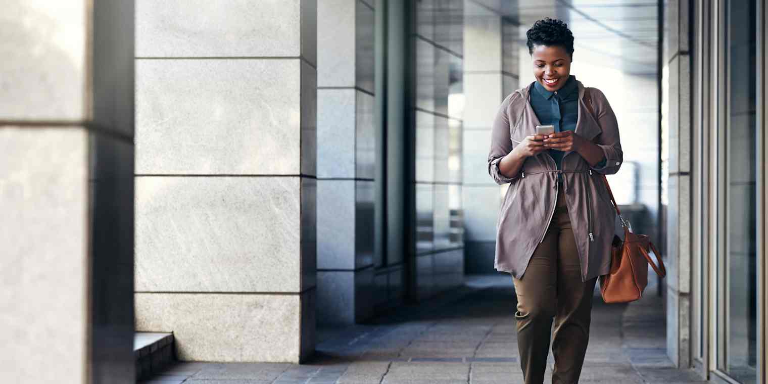 A professional woman walks through an office corridor while looking at her cell phone.