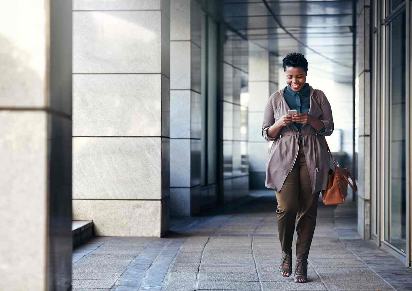 A professional woman walks through an office corridor while looking at her cell phone.