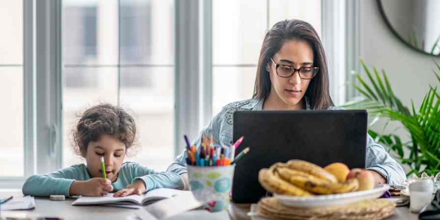 A woman works on a laptop computer at her dining room table, with a young child next to her, drawing.