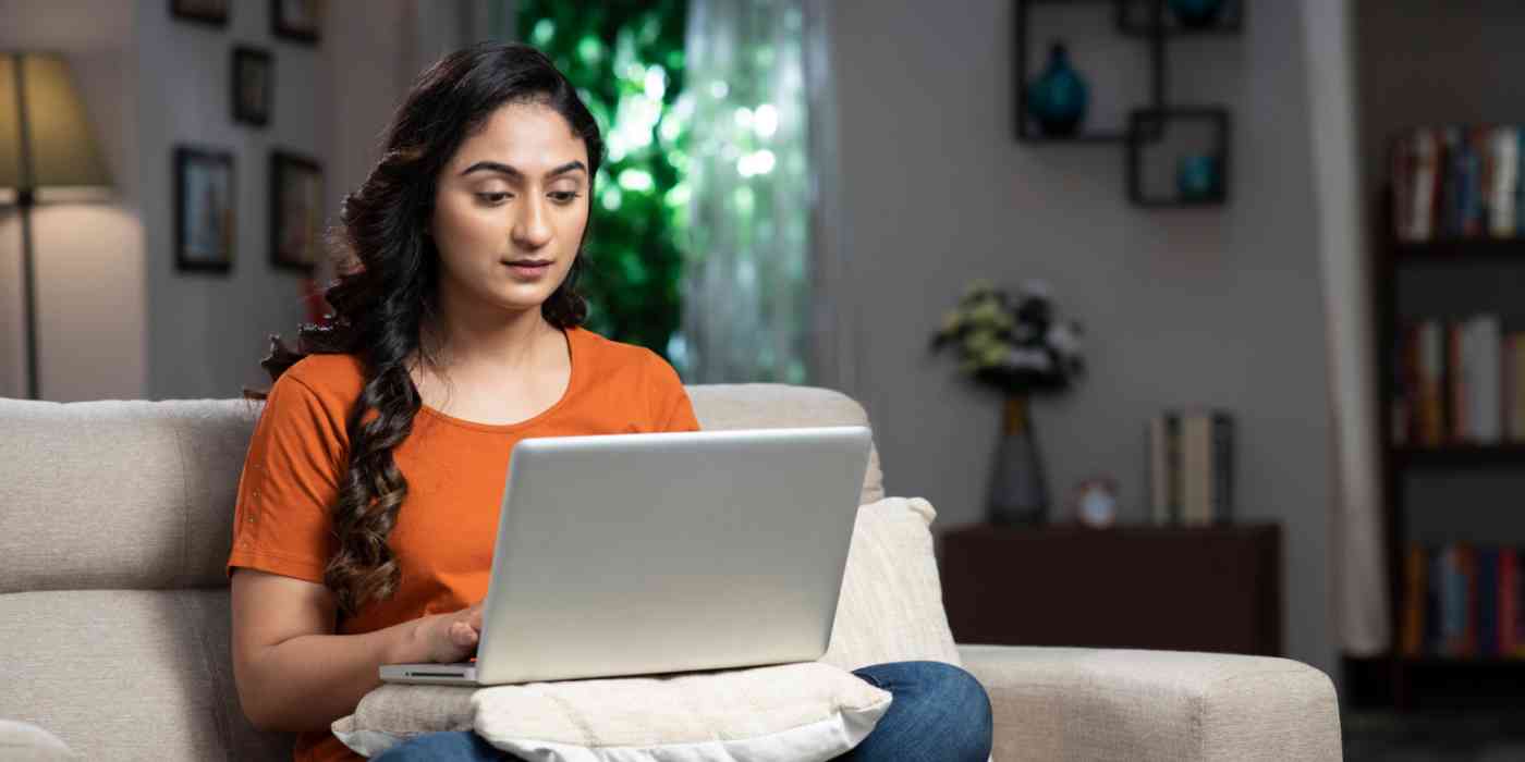 Hero image of a woman sitting on a couch with a computer on a pillow on her lap
