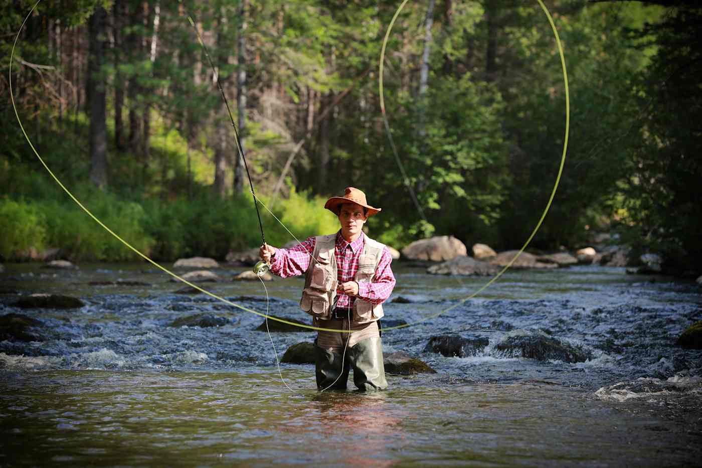 A man wears waders and a fishing vest as he stands in a river casting out his fly fishing line.