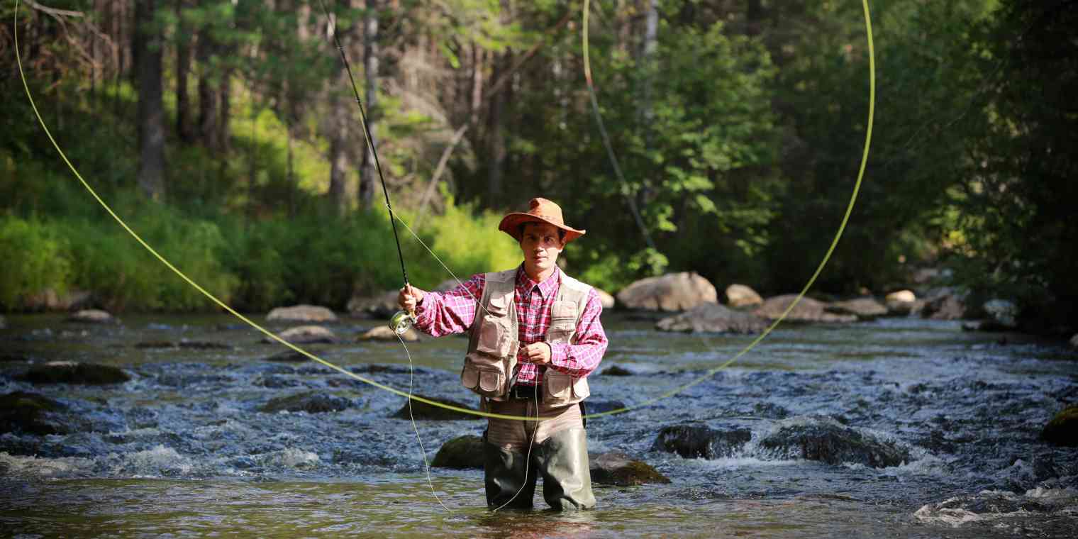 A man wears waders and a fishing vest as he stands in a river casting out his fly fishing line.