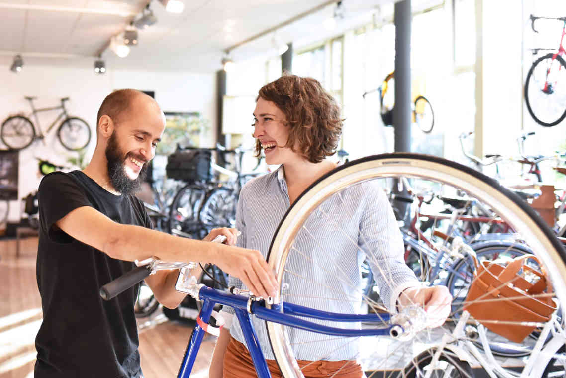 A man works on a bicycle in a bike shop while talking with a smiling customer.