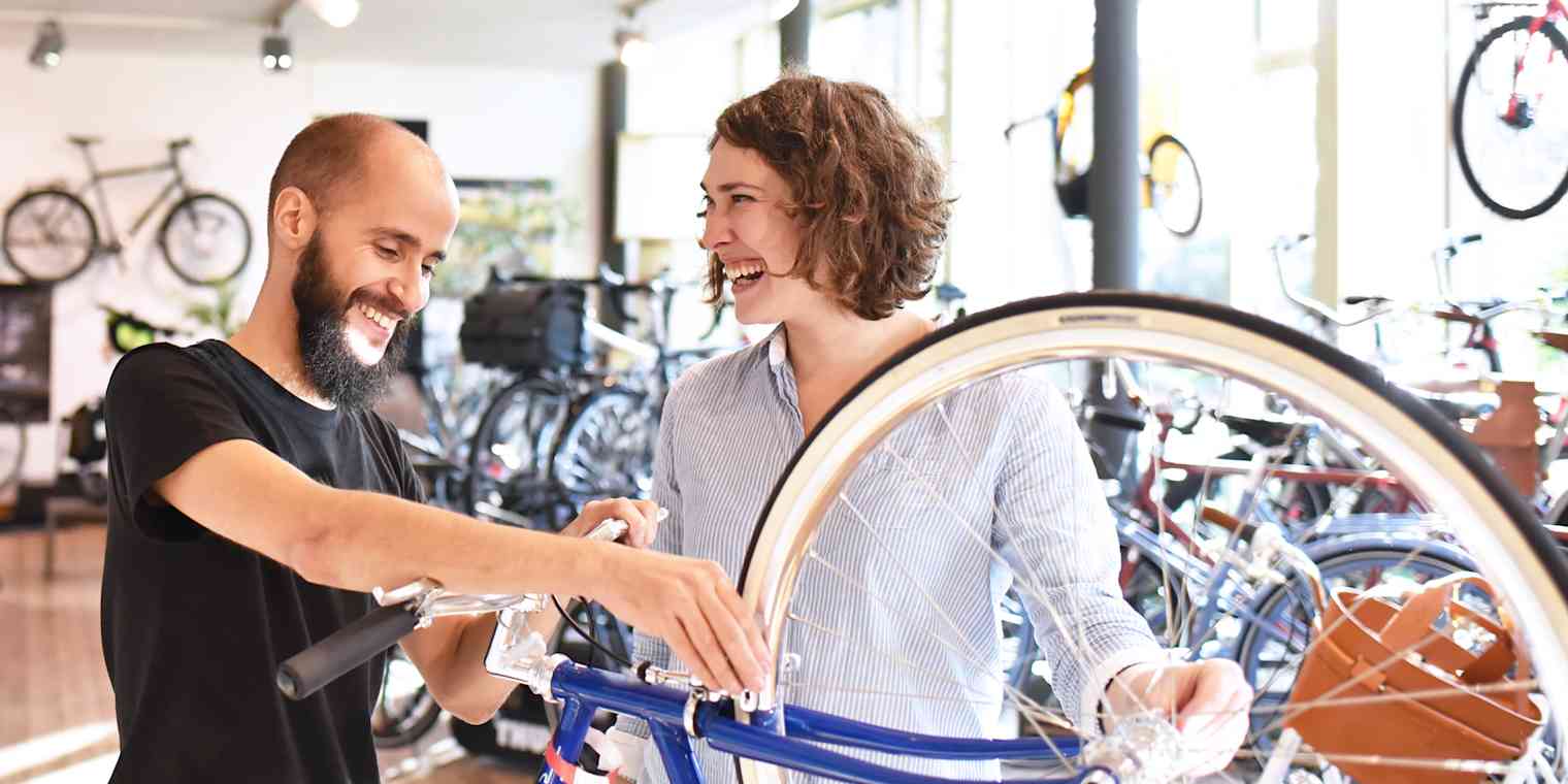 A man works on a bicycle in a bike shop while talking with a smiling customer.