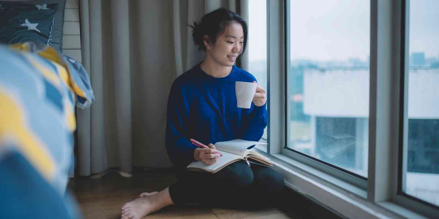 Hero image of a woman with a journal sitting next to a window, holding a mug
