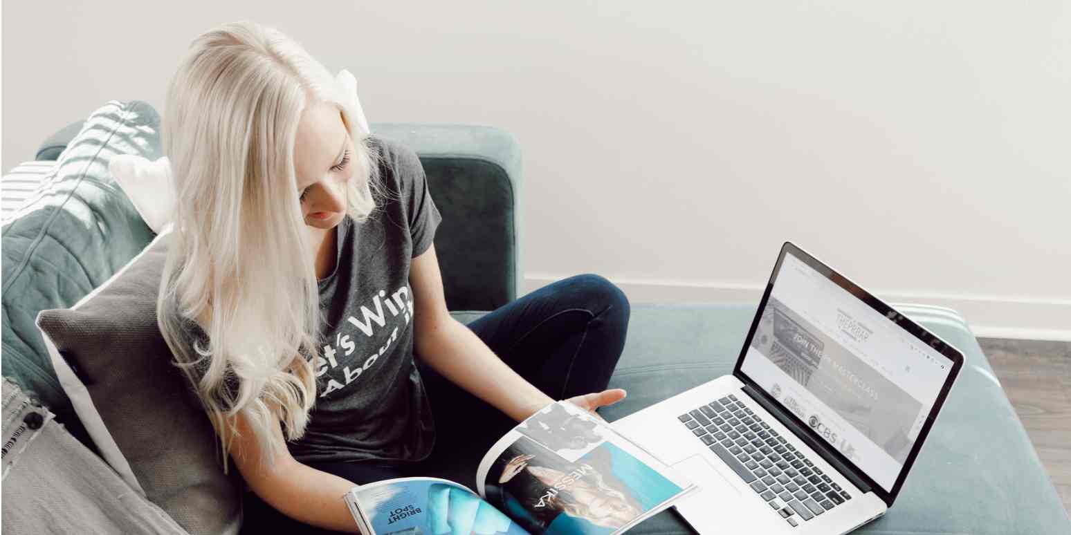 Screenshot of a woman cross-legged on a blue couch, with a laptop in front of her and flipping through a magazine