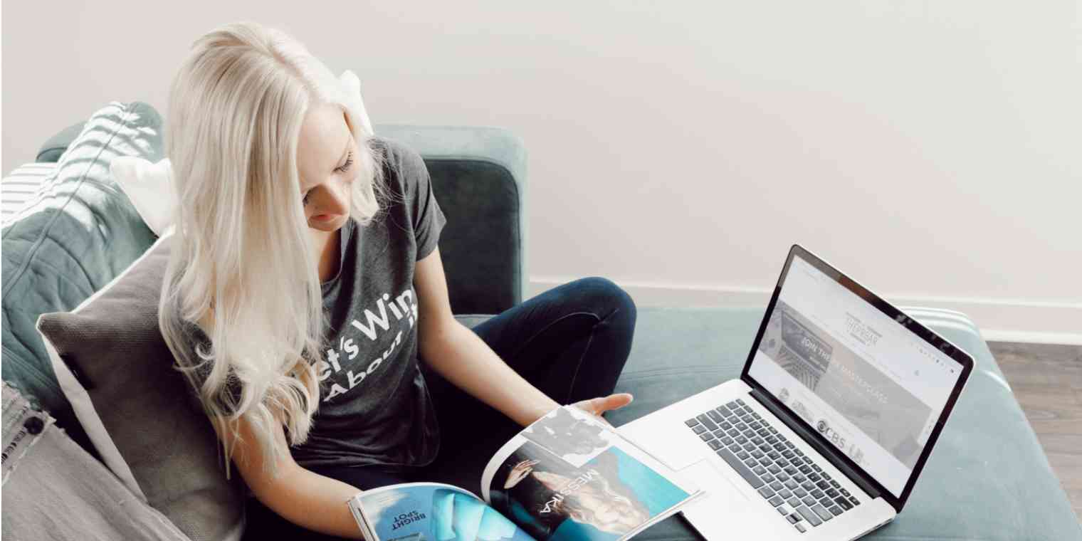 Screenshot of a woman cross-legged on a blue couch, with a laptop in front of her and flipping through a magazine