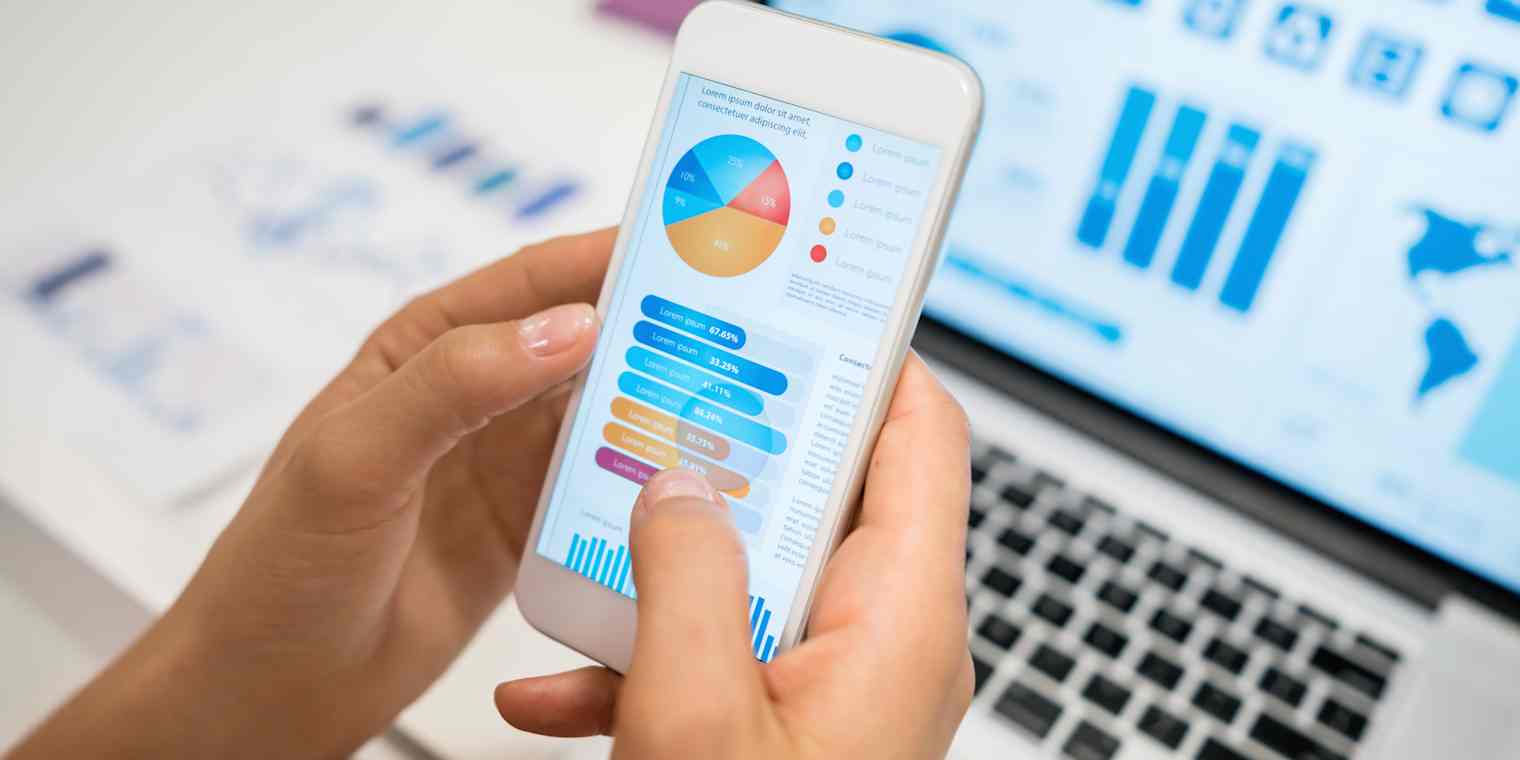 A person's hands holding a smartphone displaying charts and other information in the foreground, with a computer in the background displaying a map and charts.